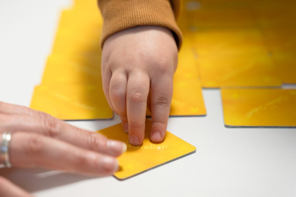 Eine Kinderhand und eine Erwachsenenhand liegen in Nahaufnahme auf der Rückseite einer gelben Memory-Karte.