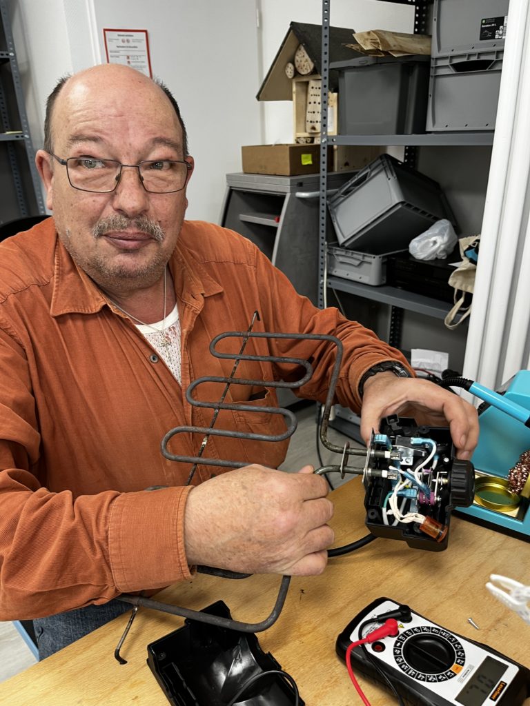 Ein älterer Mann im orangen Hemd versucht die Heizspirale eines Elektrogrills zu reparieren und blickt für das Foto in die Kamera.