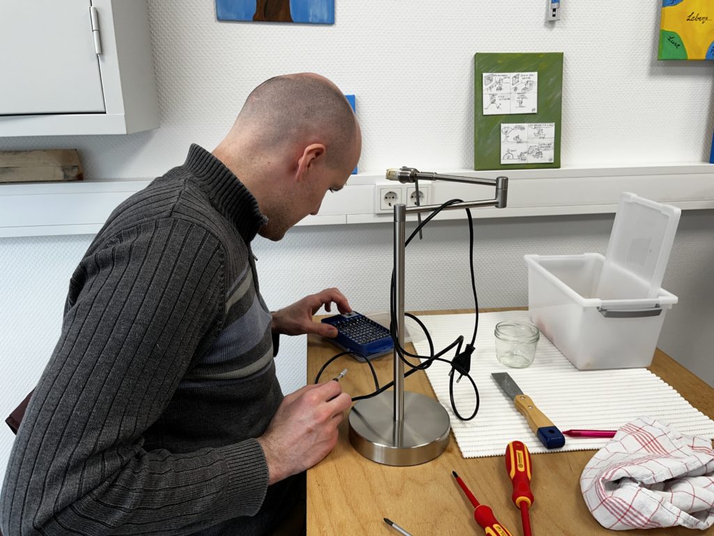 Ein jüngerer Mann sitzt an einem Tisch und begutachtet eine Lampe. Werkzeuge und Messgeräte liegen auf dem Tisch.