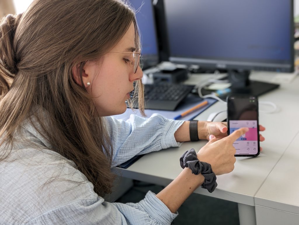 Eine junge Frau sitzt vor ihrem Schreibtisch und zeigt eine Anwendung auf ihrem Smartphone. 