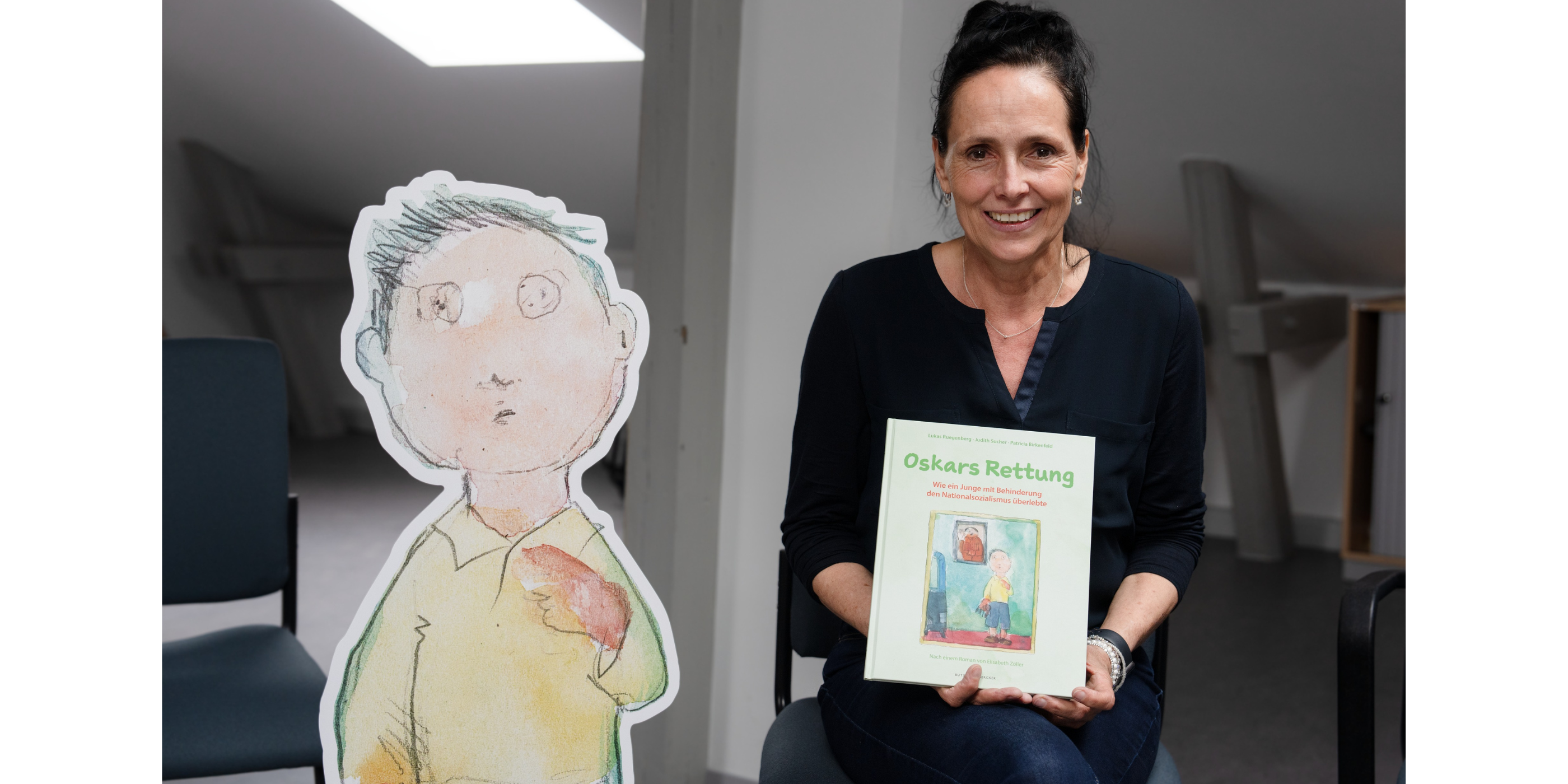 Die Autorin Patricia Birkenfeld sitzt auf einem Stuhl und hält ihr Buch lachend in die Kamera. Links von ihr steht ein Pappaufsteller der Hauptfigur Oskar.