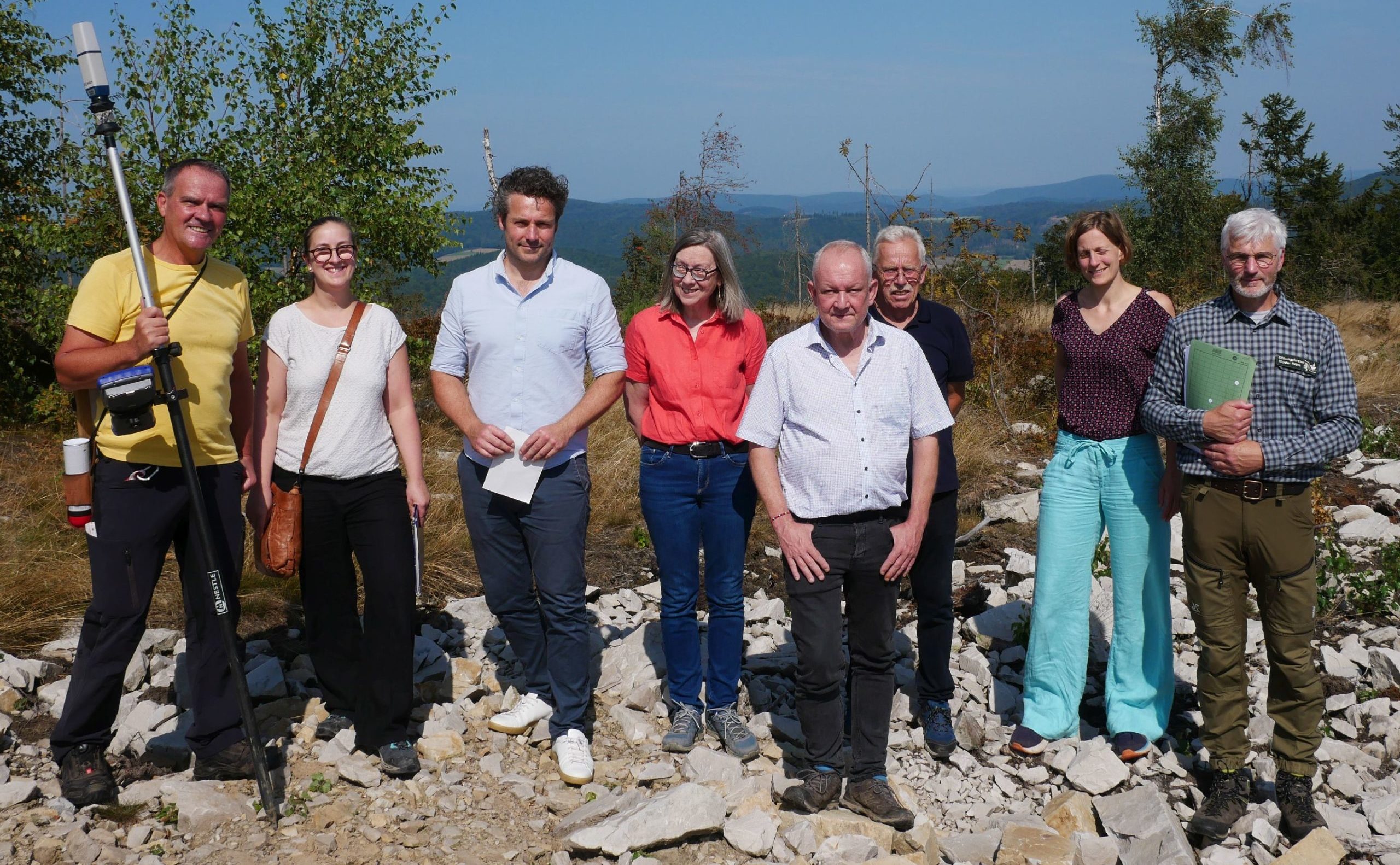 Mehrere am Bau beteiligte Personen stehen zu einem Gruppenfoto in einer Reihe. Im Hintergrund sieht man die Aussicht auf den Naturpark Kellerwald-Edersee.