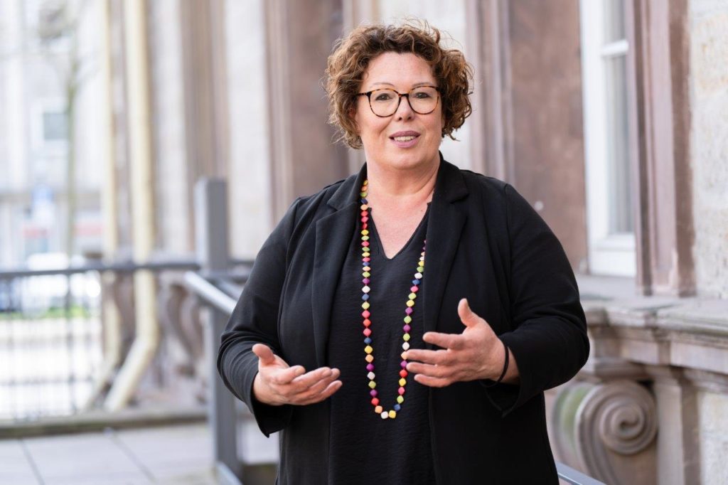 Eine Frau mit lockigen, dunklen Haaren und einer bunten Halskette schaut freundlich und einladend in die Kamera. 