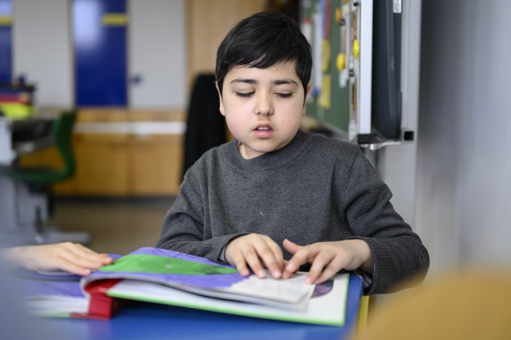 Ein Junge liest ein Buch in Brailleschrift. Neben ihm zu erkennen ist die grüne Schultafel.