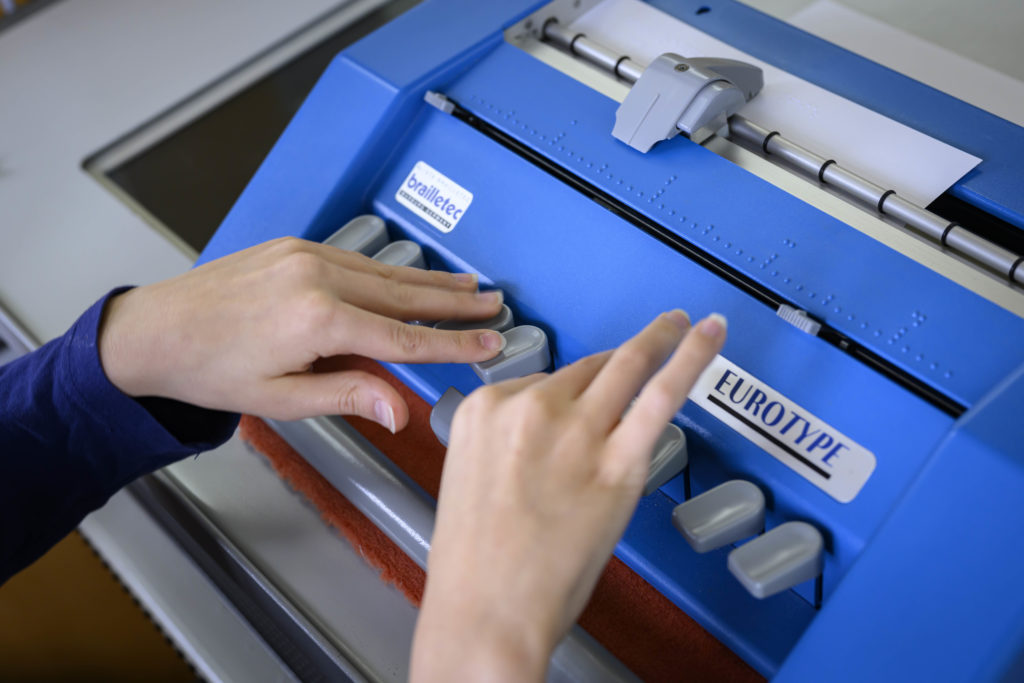 Eine Nahaufnahme der blauen Blindenschreibmaschine. Jemand tippt darauf. Deutlich zu erkennen ist die Brailleschrift an den Tasten.