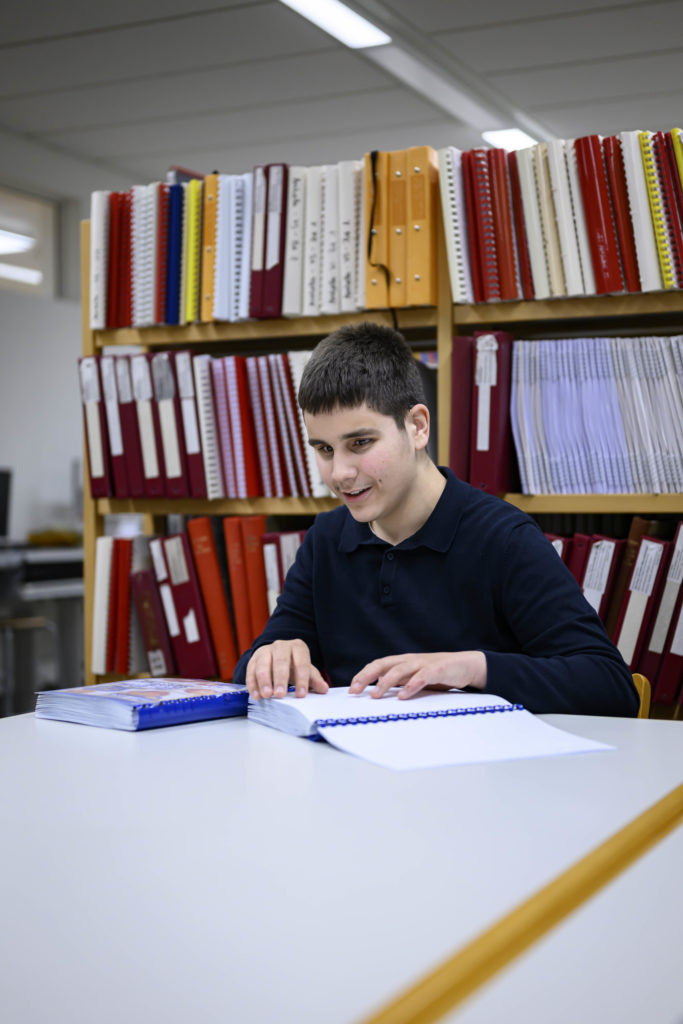 Ein Junge mit schwarzem Haar fährt mit den Fingern über ein Buch in Brailleschrift. Im Hintergrund erkennt man mehrere rote Orner in einem Regal.