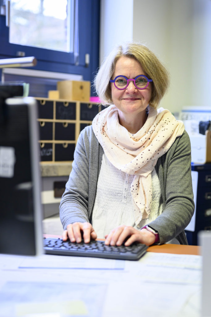 Eine Frau mit einer auffälligen lila-blauen Brille sitzt lächelnd an ihrem Computer.