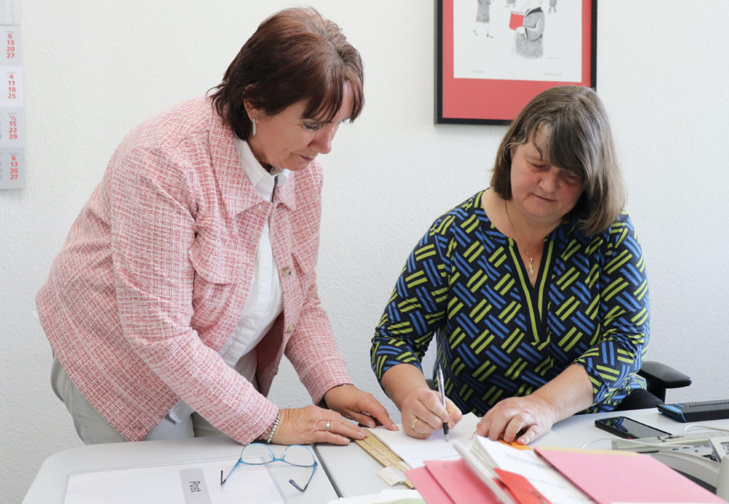 Zwei Frauen in einer Büroümgebung. Die eine, sitzende Frau, hält in der rechten Hand deutlich erkennbar einen Stift.