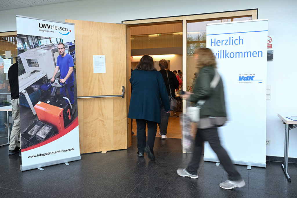 Personen gehen durch die geöffnete Tür in den Saal, rechts und links sind Roll-Ups von Integrationsamt und VdK