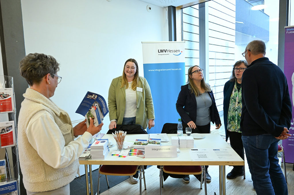 Zwei Besucher stehen mit Info-Blättern vor dem Stand des LWV Hessen Integrationsamtes, dahinter drei Mitarbeiterinnen