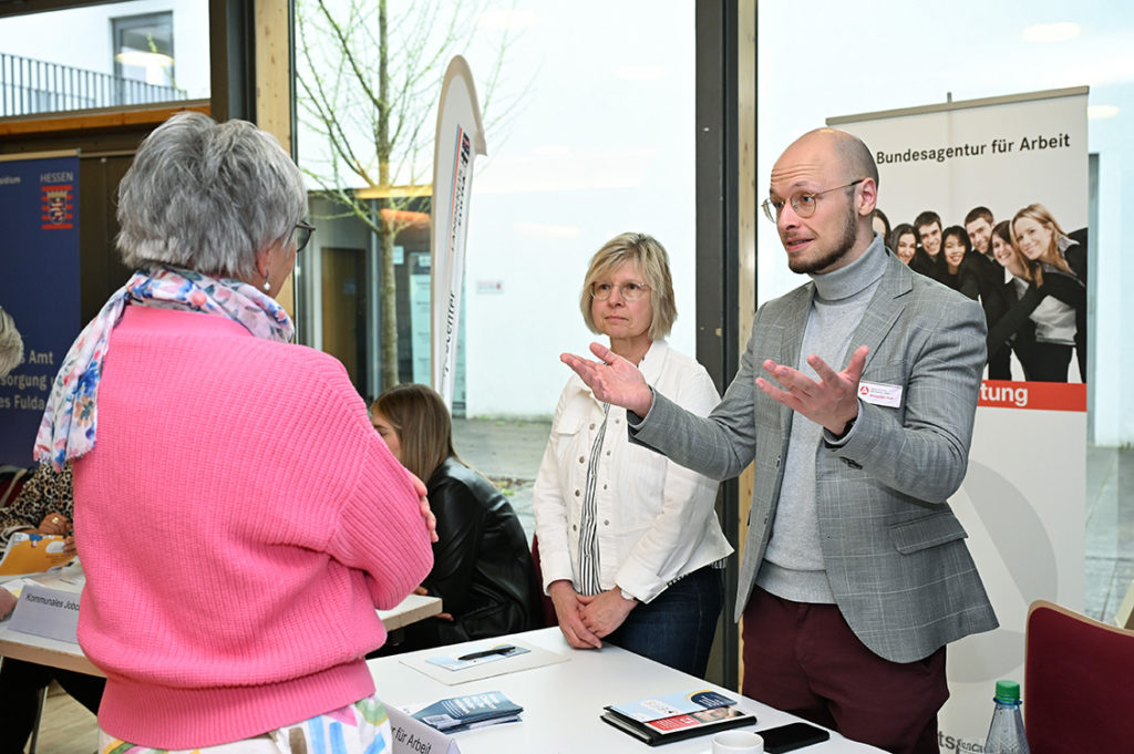 Eine Frau steht vor dem Stand der Bundesagentur für Arbeit, ein Mann erklärt ihr etwas mit ausgebreiteten Armen