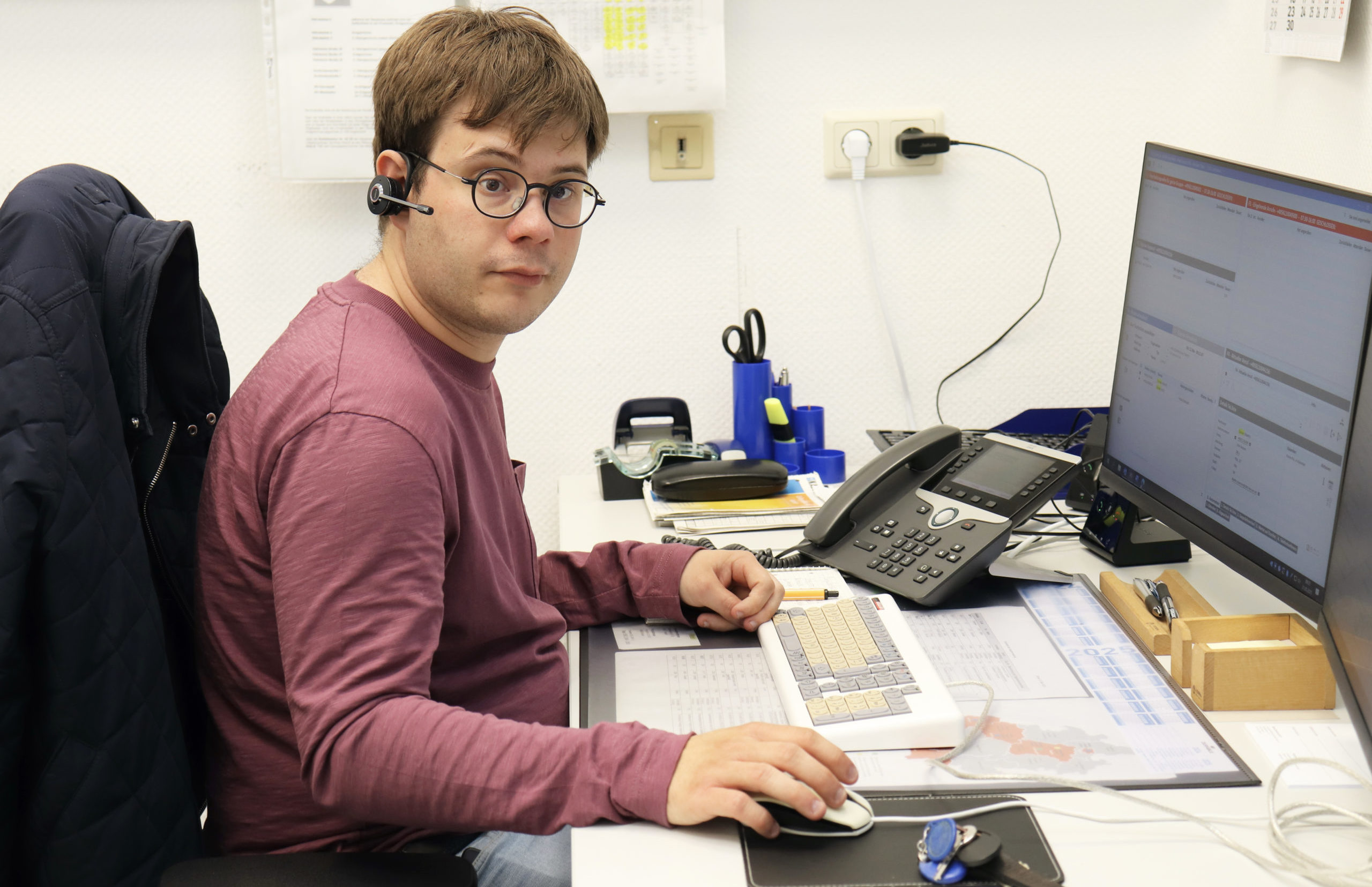 Ein Mann mit rotem Pullover und Brille sitzt an einem Büroarbeitsplatz. Er trägt ein Headset und links von ihm auf dem Tisch steht sein Telefon.