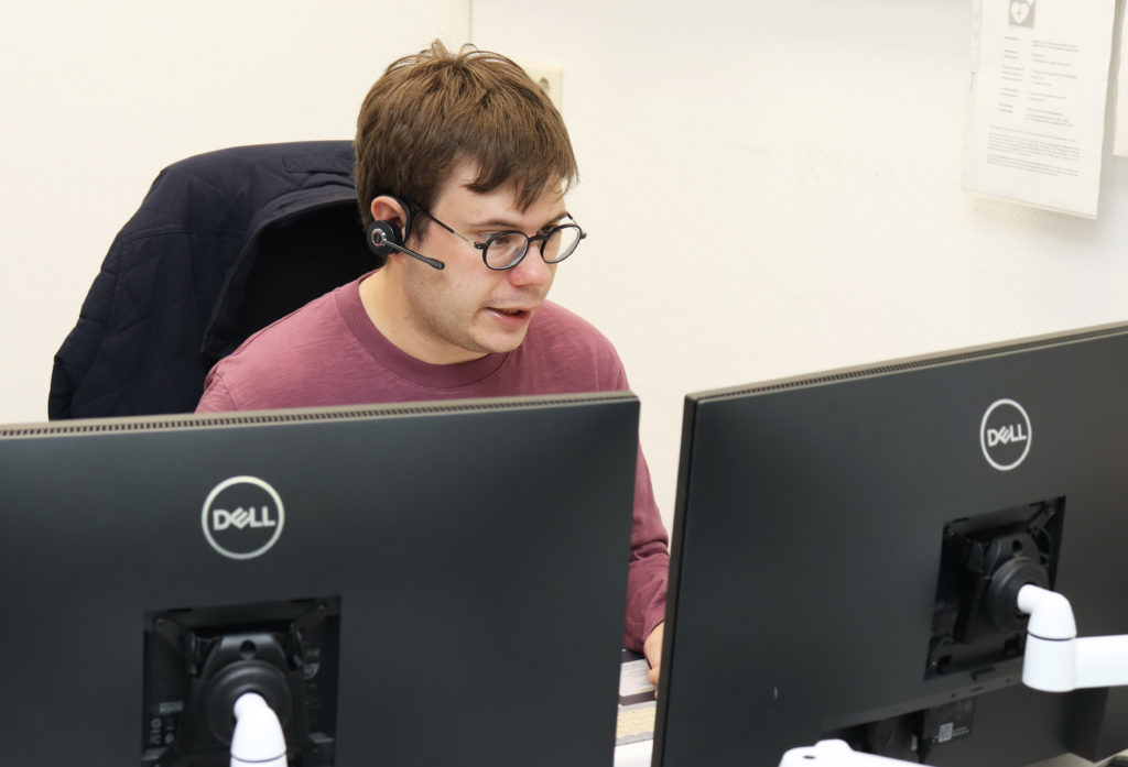 Ein Mann vor seinen zwei Bildschirmen. Deutlich zu erkennen, sein Headset am rechten Ohr und die zwei Computermonitore im Vordergrund.