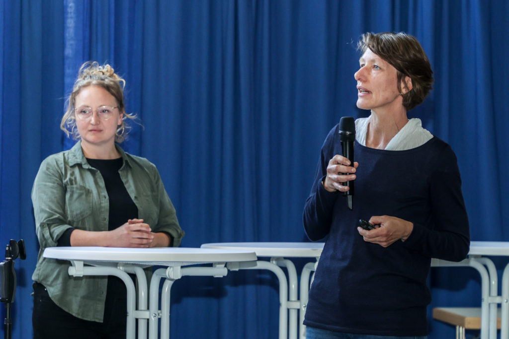 Eine Frau mittleren Alters mit einem dunkelblauen Pullover spricht durch ein Mikro und hält in der anderen Hand einen Presenter zur Steuerung der Präsentation.