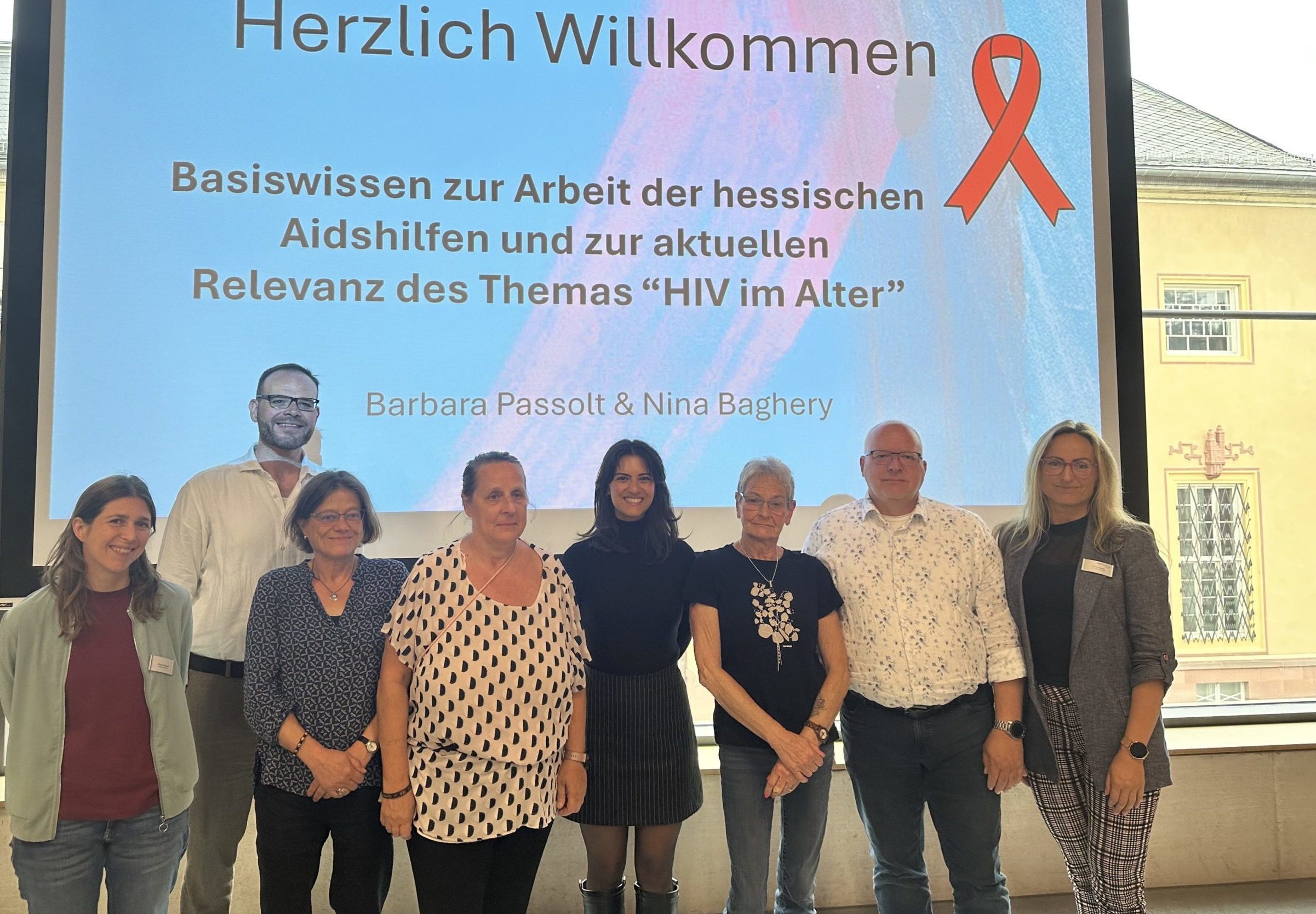 Die Beteiligten der Veranstaltung haben sich zu einem Gruppenfoto vor der eingeschalteten Leinwand versammelt und lächeln in die Kamera.