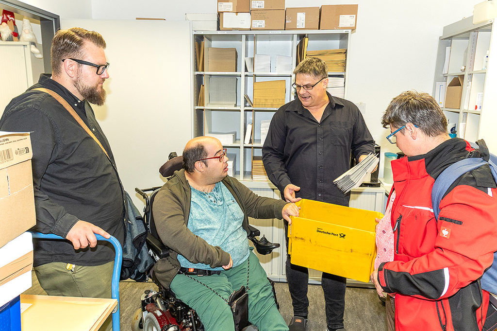 Ein Mann mit einem gelben Plastikkarton. Das Skript man mit einem gelben Plastikkarton und einem Stapelbriefen in der Hand erklärt einer Gruppe von Menschen etwas.