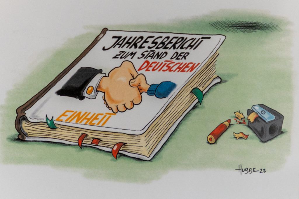 Ein Cartoon zeigt ein Buch mit der Aufschrift "Jahresbericht zum Stand der deutschen Einheit". Man sieht einen Handschlag einer sehr großen und sehr kleinen Hand darauf.