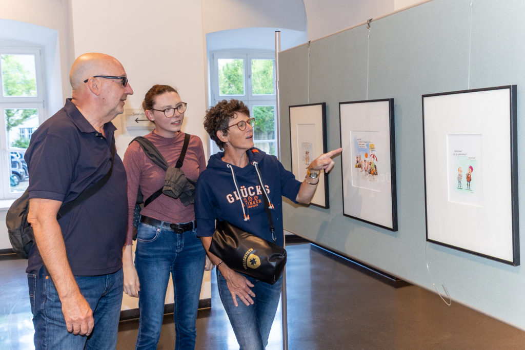 Drei Besucher der Ausstellung deuten belustigt auf die aufgehängten Cartoons.