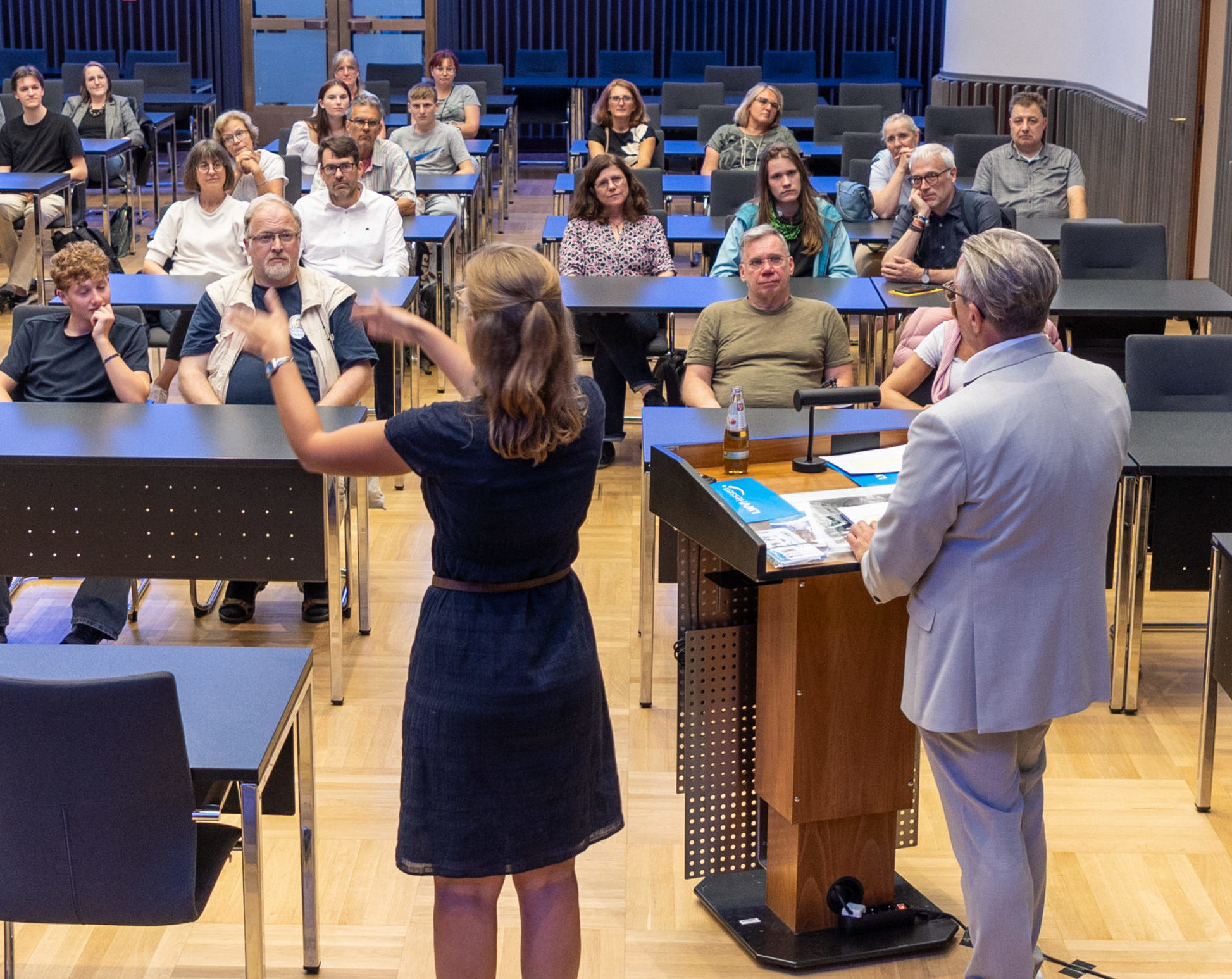 Die Menschen sitzen in Tischreihen in einem Saal, während der Führung. Im Vordergrund gestikuliert die Dolmetscherin.