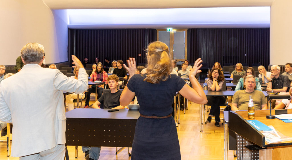 Die Menschen sitzen in Tischreihen in einem Saal, während der Führung. Im Vordergrund gestikuliert die Dolmetscherin.