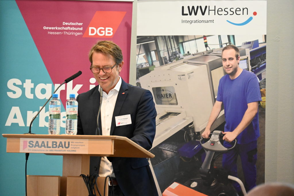 Ein Mann um die 50 mit Brille und schwarzem Anzug lacht während seines Vortrags auf der Bühne herzlich.