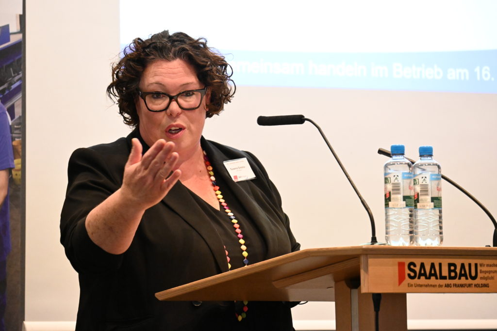 Eine Frau mit schwarzen Haaren und schwarzer Bluse hält auf der Veranstaltung einen Vortrag und gestikuliert.