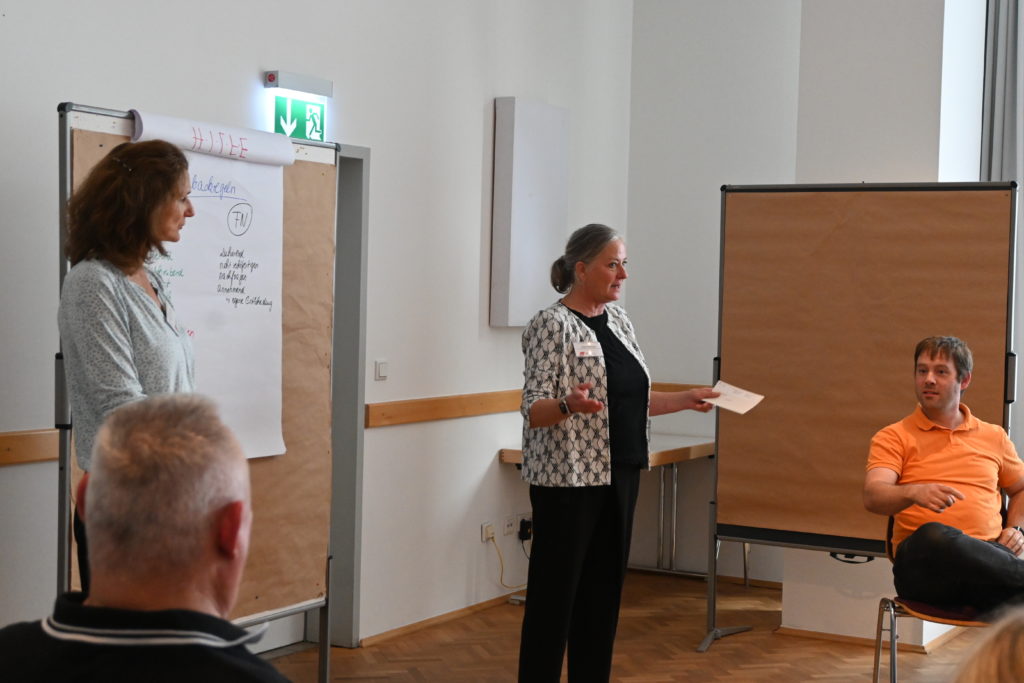Auf dem Bild sieht man eine Frau mit ausgebreiteten Armen und Moderationskarten in der linken Hand. Im Hintergrund sieht man einen Flipchart.