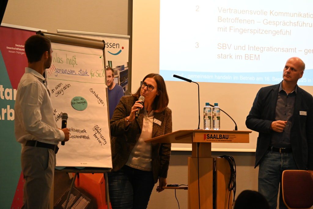 Drei Personen diskutieren auf der Veranstaltung an einem Flipchart. Im Hintergrund sieht man eine aufgeblendete PowerPoint-Präsentation.