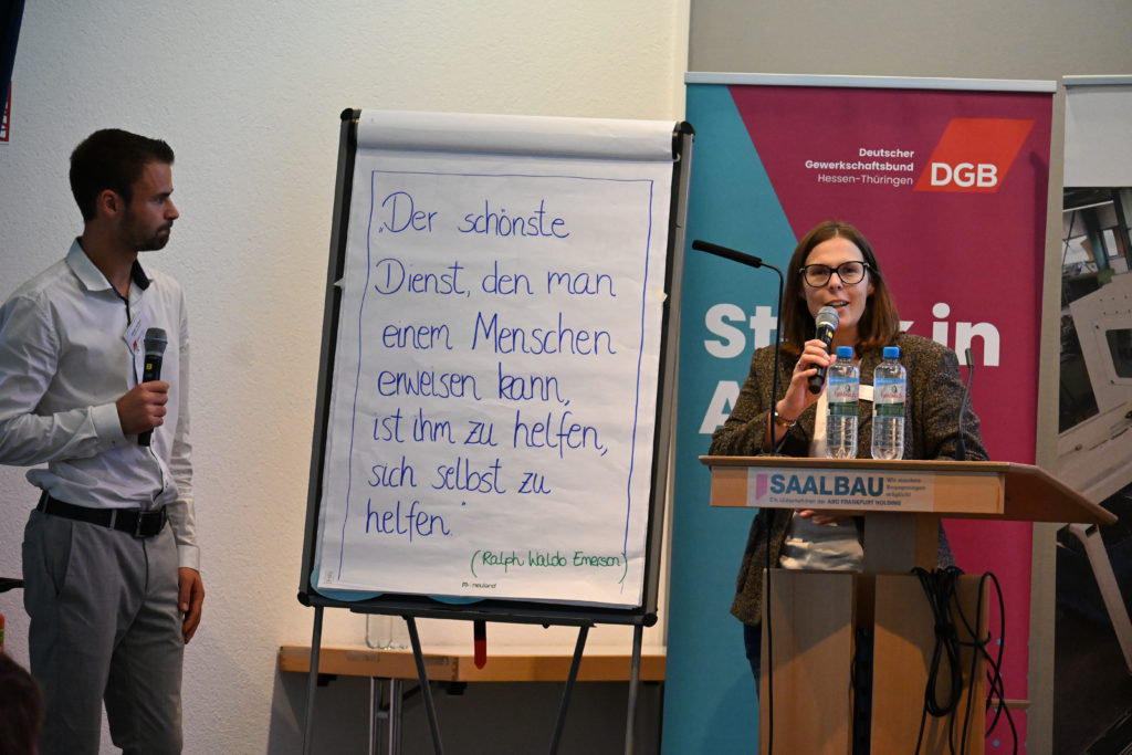 Eine Frau und ein Mann stehen mit Mikrofon in der Hand an einem Flipchart, der mit großen blauen Buchstaben beschriftet ist.