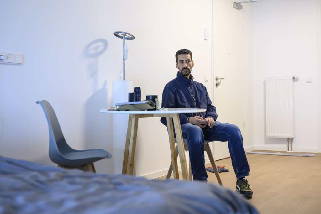 Ein Mann in einer blauen Jacke sitzt auf einem Holzstuhl in einer hellen Wohnung.