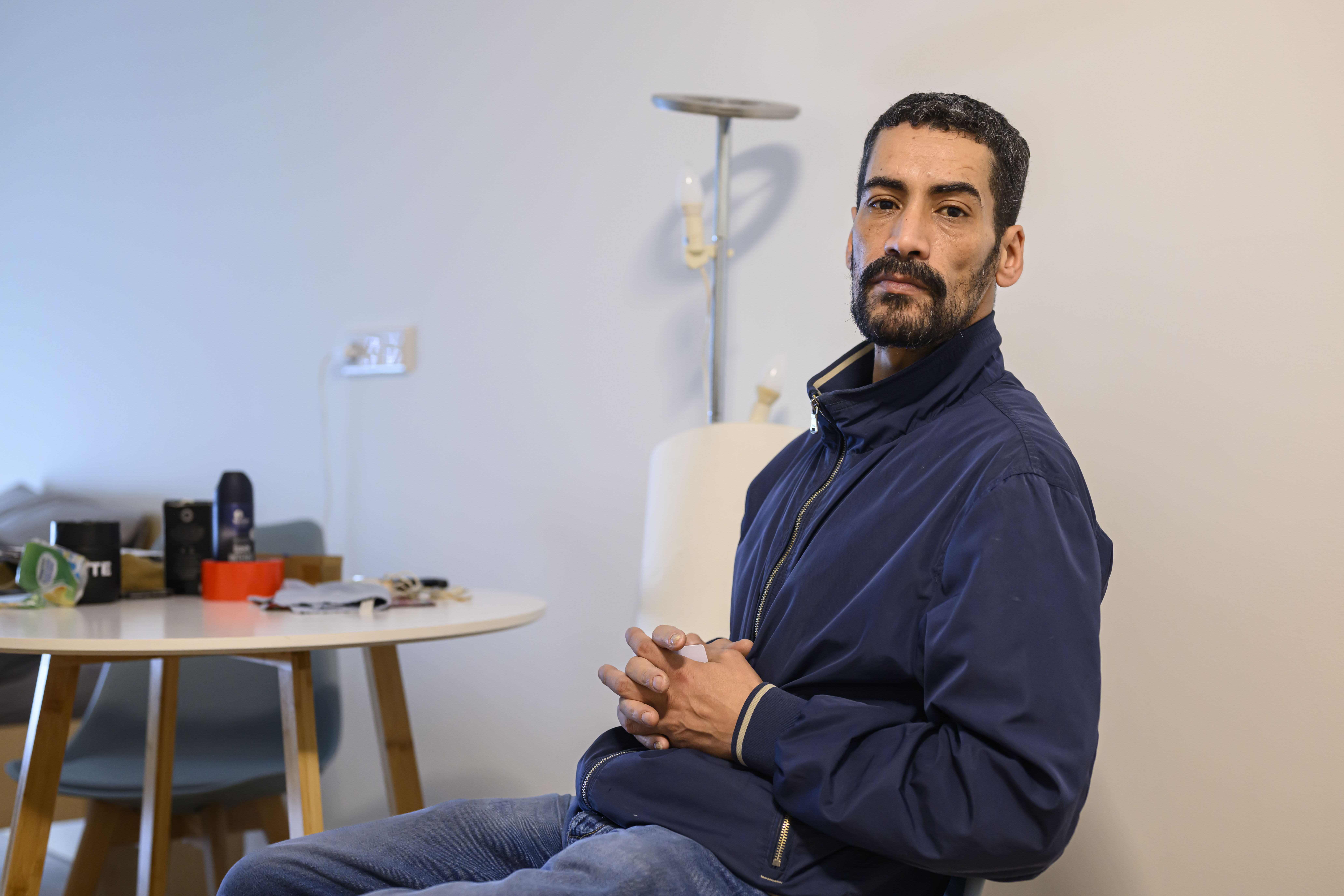 Endlich ein Zimmer für sich allein. Walid Mejri ist nach längerer Zeit der Wohnungslosigkeit wieder zufrieden. Ein Mann mit einer blauen Jacke sitzt mit gefalteten Händen auf einem Stuhl und schaut ernst in die Kamera.