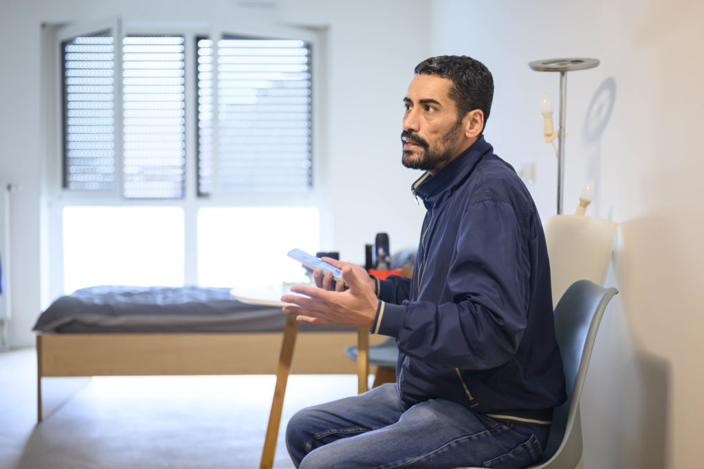 Der Mann mit der blauen Jacke sitzt auf einem Stuhl in seiner Wohnung und erklärt etwas. In der rechten Hand hält er ein Smartphone.