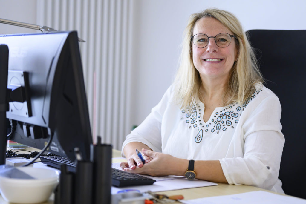 Eine Frau in einer weißen Bluse sitzt am Schreibtisch hinter dem Computerbildschirm und lächelt in die Kamera.