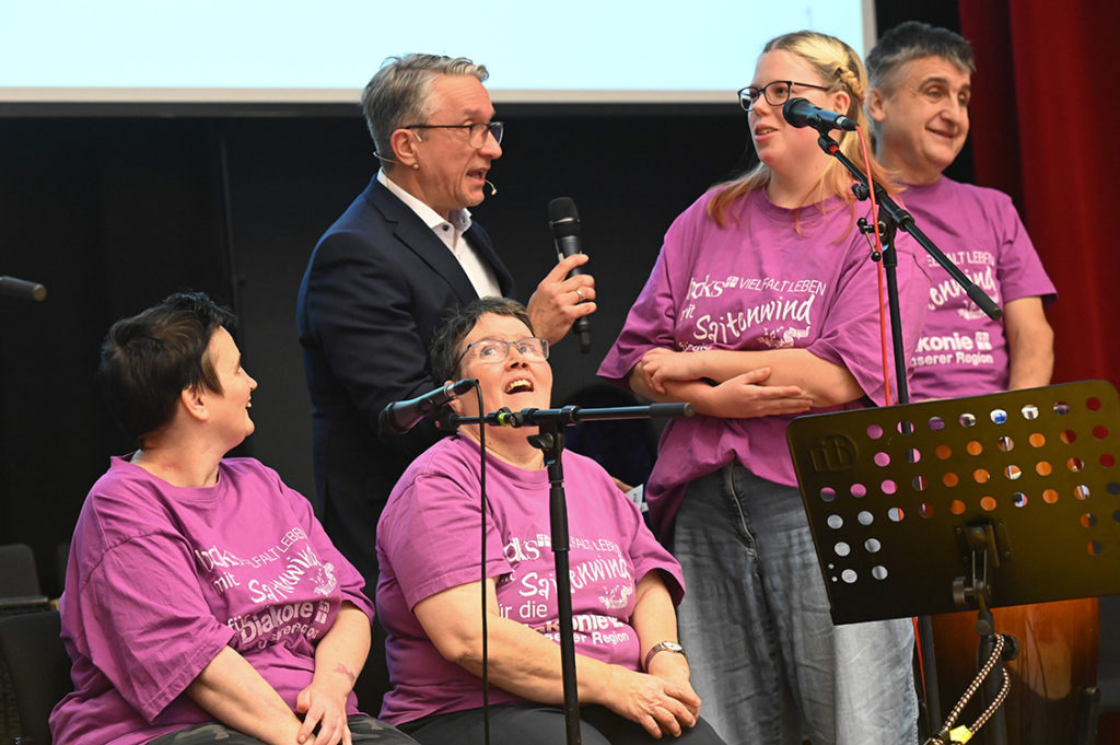 Ein Moderaor zwei Mitglieder der Band. Die anderen Bandmitglieder schauen interessiert zu.