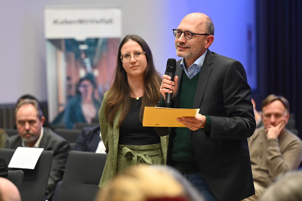 Ein Mann stellt nach dem Vortrag eine Frage über das Mikro. In der linken Hand hält er mehrere Blatt Papier.