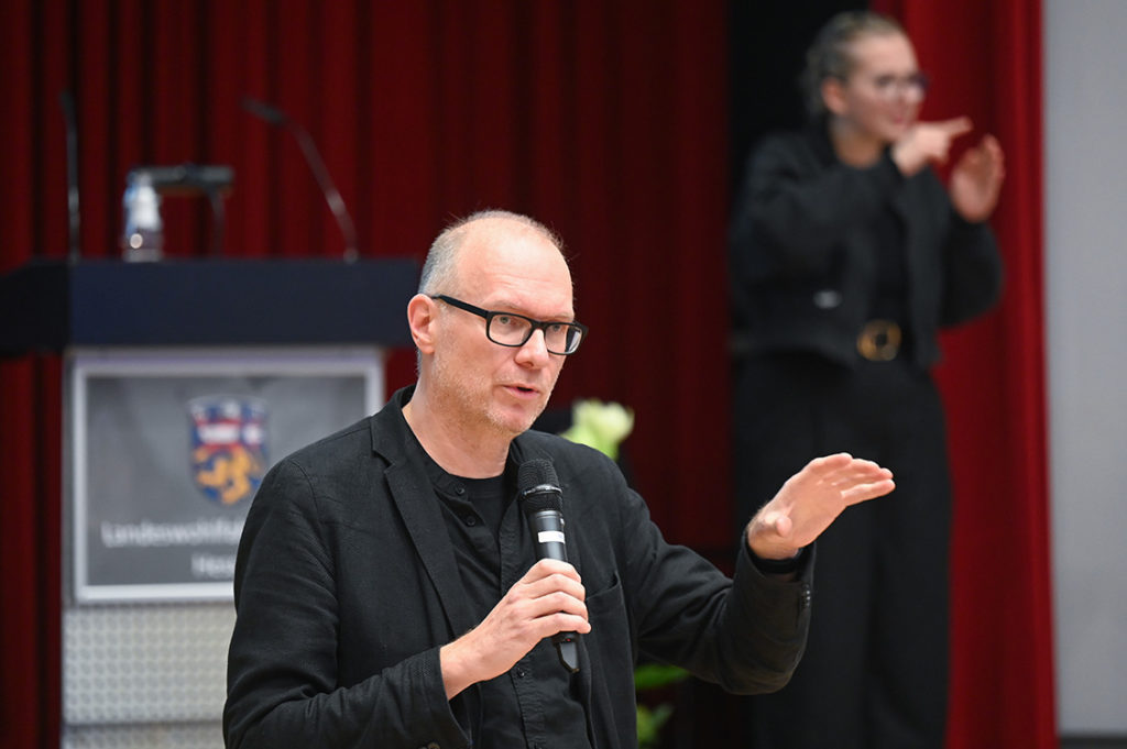 Der Mann mit der schwarzen Brille hält das Handmikro in der rechten Hand und gestikuliert mit der linken Hand.