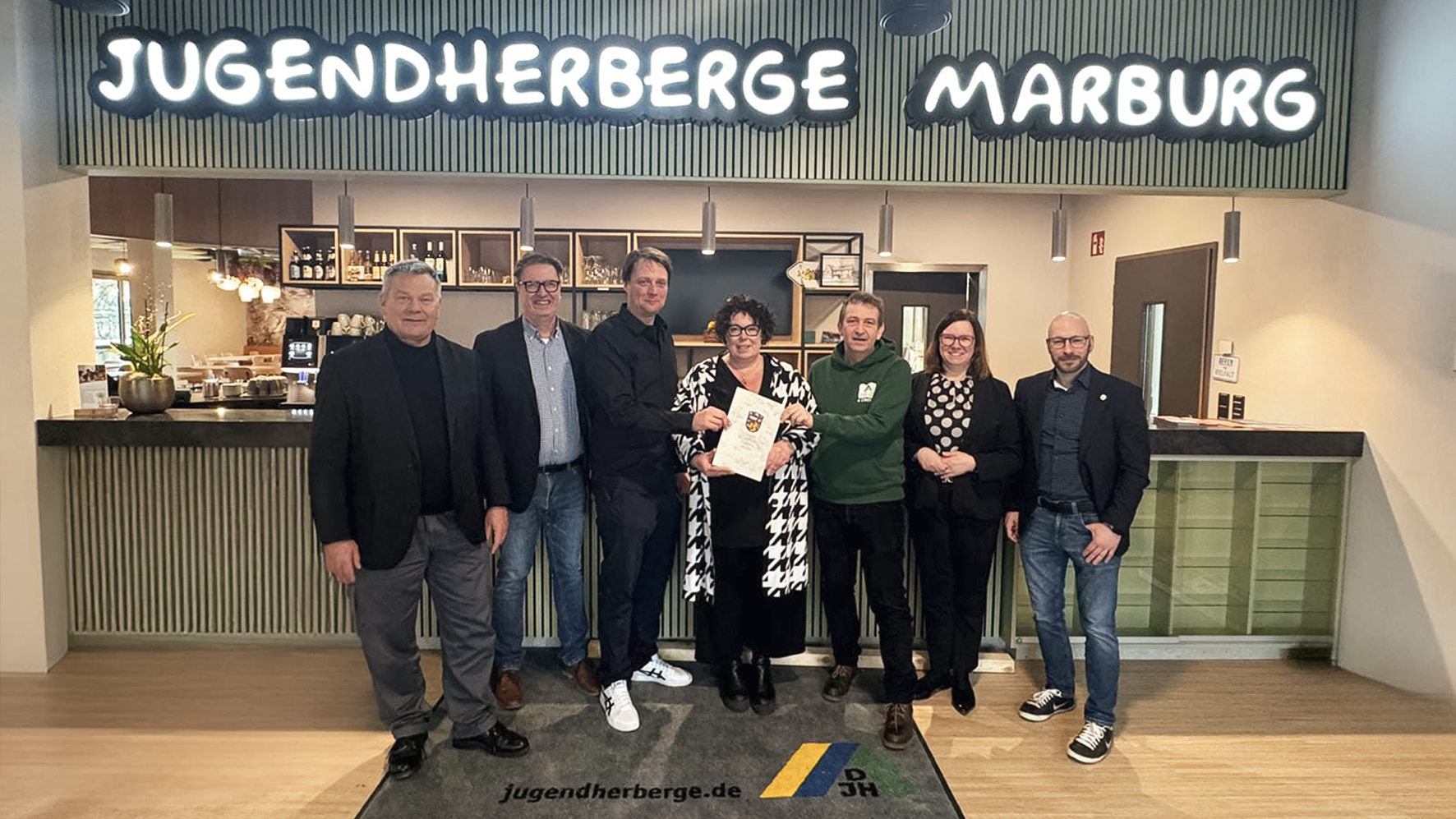 Sieben erwachsene stehen vor der Rezeption der Jugendherberge Marburg in einer Reihe (v.l): Dr. Thomas Spies, Oberbürgermeister von Marburg; Dr. Klaus Becker und Knut Stolle, beide Vorstand DJH Hessen e.V.; Susanne Simmler, LWV-Landesdirektorin; Peter Schmidt, Leiter der Jugendherberge; Manuela Strube, Staatssekretärin im Hessischen Sozialministerium und Sebastian Sack, Abgeordneter im Hessischen Landtag.
Die Frau in der Mitte hält den Bescheid mit dem Wappen des Landes Hessens in der Hand.
Über den Personen an der Wand hängt in Leuchtschrift: "Jugendherberge Marburg".