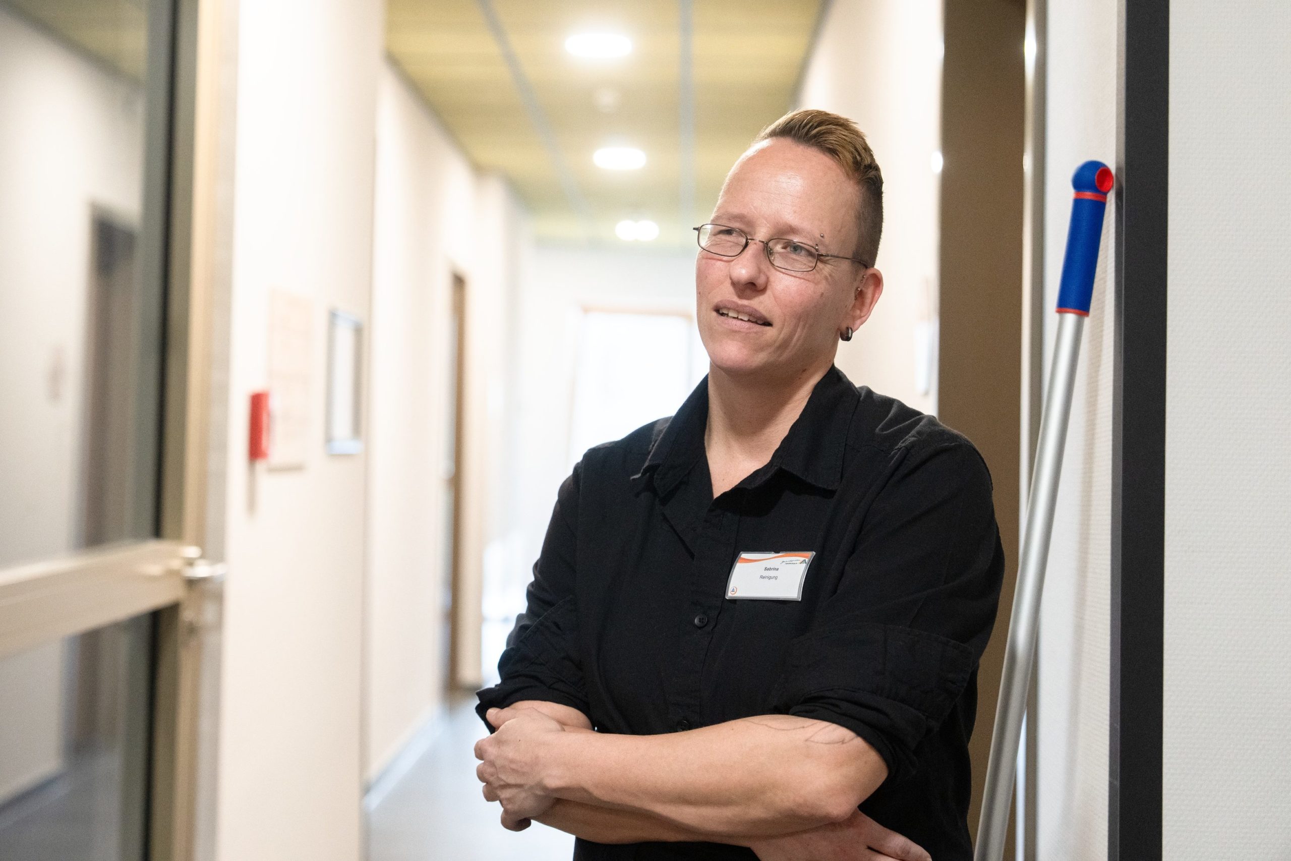 Eine Frau lehnt an der Wand im Gang einer Jugendherberge. Sie trägt ein schwarzes T-Shirt mit Namensschild.