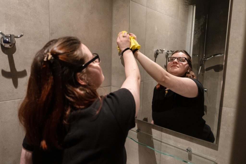 Eine junge Frau wischt mit einem gelben Tuch über einen Spiegel. Sie trägt ein schwarzes Tshirt und eine schwarze Sonnenbrille. Man sieht sie von hinten und ihre Spiegelung von vorne im Spiegel.