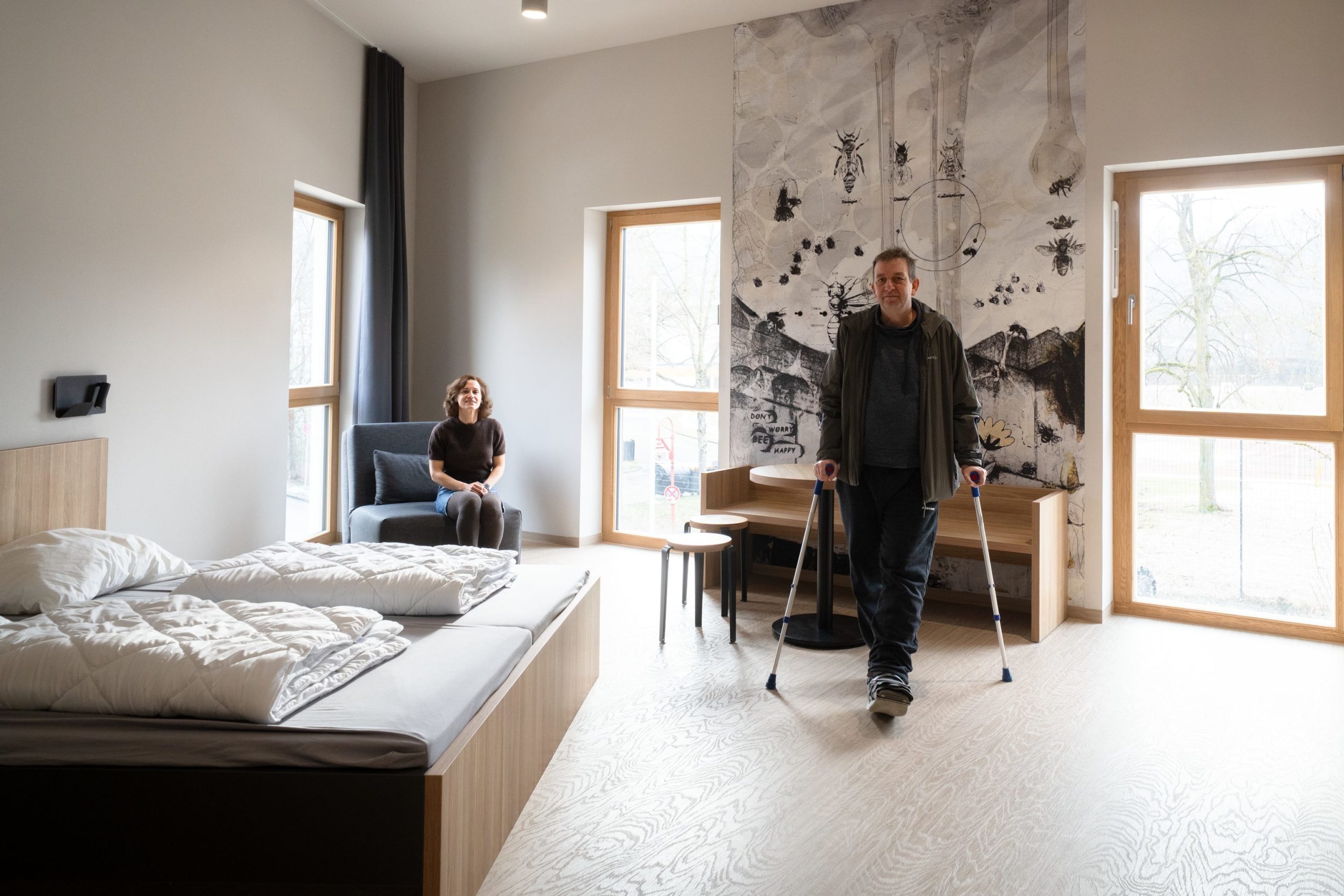 Ein barrierefreies Zimmer in einer Jugendherberge: Ausreichend Platz zum Bewegen, ein barrierefreies Bett, eine Frau sitzt auf einem blauen Sessel in einer Zimmerecke und schaut in die Kamera, ein Mann steht auf Gehhilfen mitten im Raum. Bodentiefe Fenster erhellen den Raum.