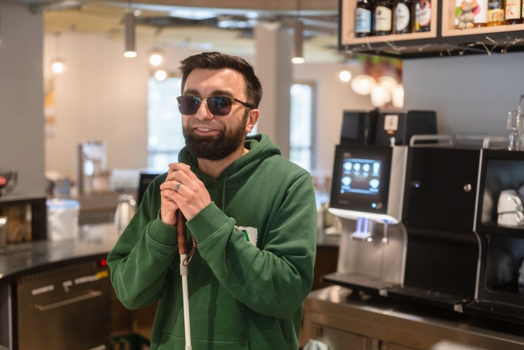 Ein Mann steht vor einem Kaffeevollautomaten hinter einer Rezeption. Er trägt einen grünen Pulli sowie eine dunkle Sonnnbrille und stützt sich auf seinen Blindenstock.