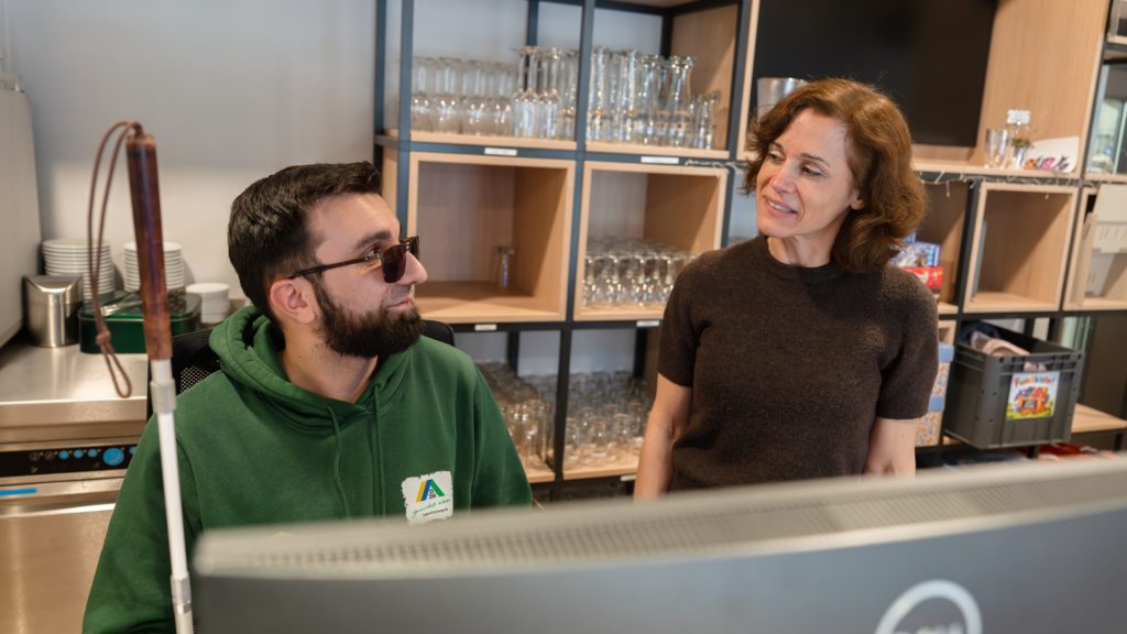 Zwei Personen hinter der Rezeption schauen sich an. Der Mann links trägt einen grünen Pulli mit dem Logo der Deutschen Jugendherbergen und eine dunkle Sonnenbrille. Die Frau rechts neben ihm trägt ein schwarzes Tshirt und braune schulterlange Haare.