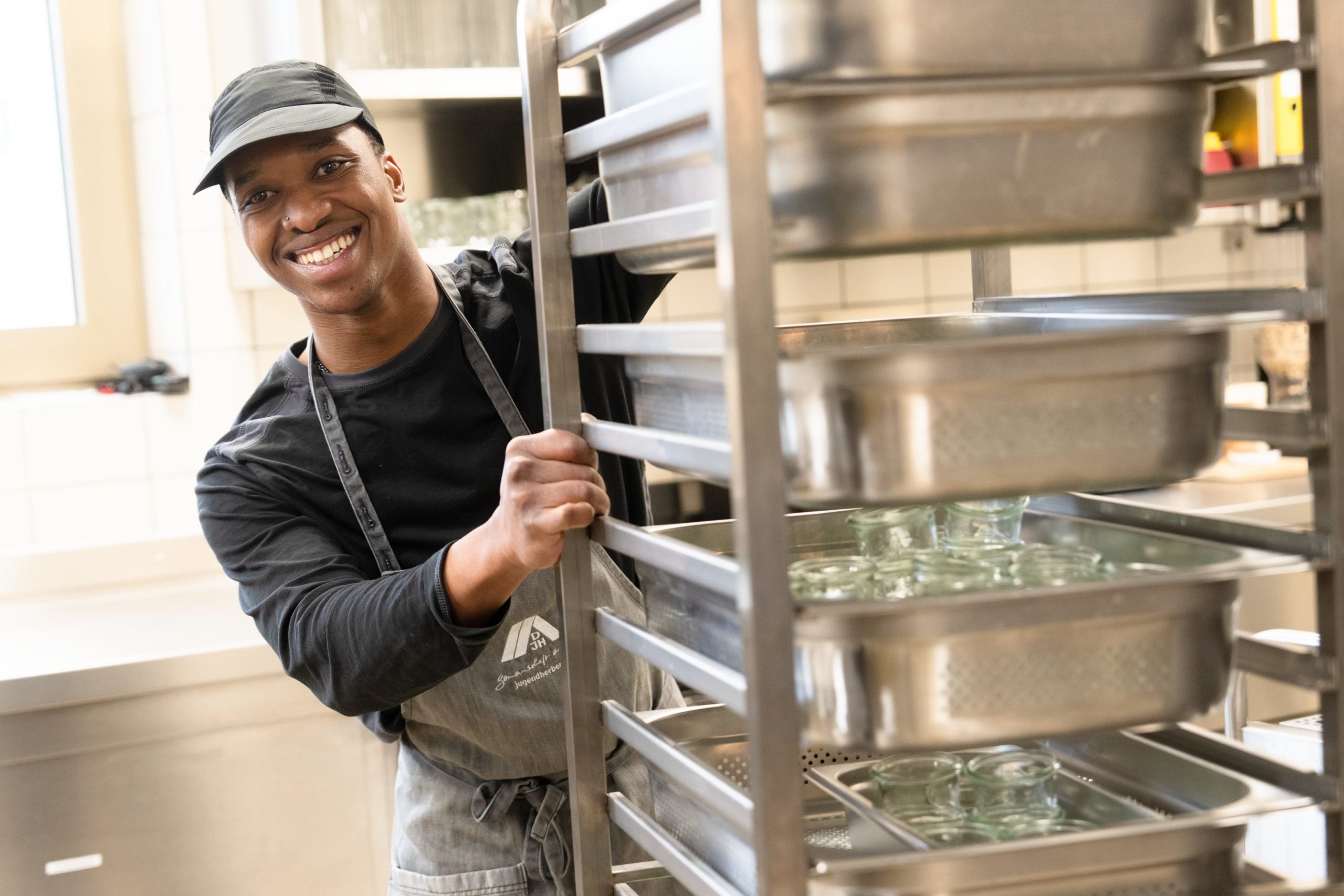 Ein Mann schiebt einen hohen Wagen mit Gläsern in einer Großküche. Er schaut links hinter dem Wagen hervor und lächelt in die Kamera.