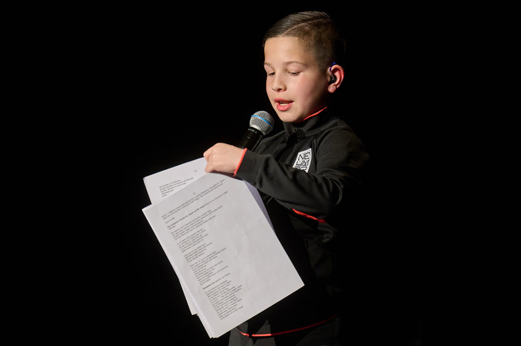 Ein Junge mit blonden Haaren steht am Mikrofon. In der Hand hält er bedrucktes Papier. 