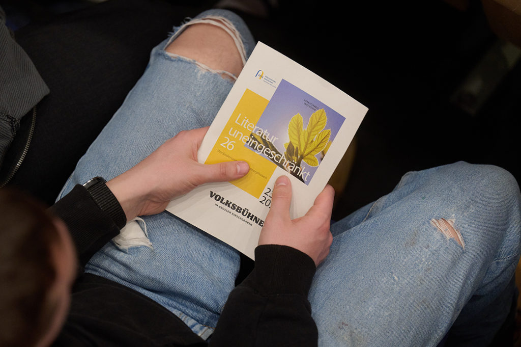 Fotografie von oben auf die Knie eines Besuchers. In beiden Händen hält er das Programmheft der Veranstaltung.