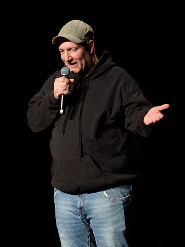 Ein Mann mit einer braunen Schirmmütze hält ein Handmikrofon in der einen Hand und macht mit der anderen Hand eine einladende Geste. 