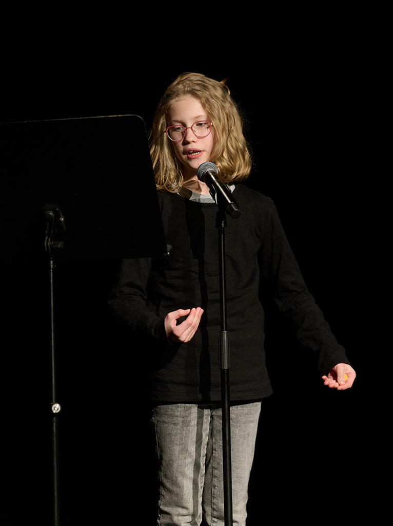 Ein Mädchen mit blonden Haaren und einer runden Brille steht am Mikrofonständer, vor schwarzem Hintergrund.