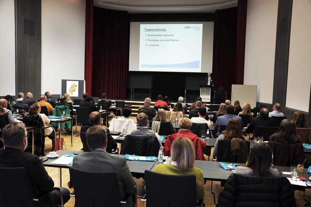 Viele Menschen im Saal hören einem Mann zu, der auf einer Bühne am Rednerpult steht.