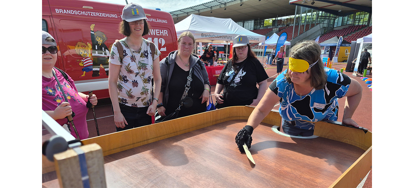 LWV-Mitarbeiterin Birgit Riester demonstrierte vorm LWV-Infostand, wie ihre Sportart Tischball/Showdown gespielt wird. Viele Gäste des inklusiven Sportfestes waren interessiert und probierten auch selbst mal aus. (alle Fotos: LWV-Öffentlichkeitsarbeit) Vier Frauen schauen auf eine Showdown-Spielplatte,. LWV-Mitarnbeiterin Birgit Riester hat einen flachen Schläger in der Hand und eine Brille auf und zeigt, wie gespielt wird.